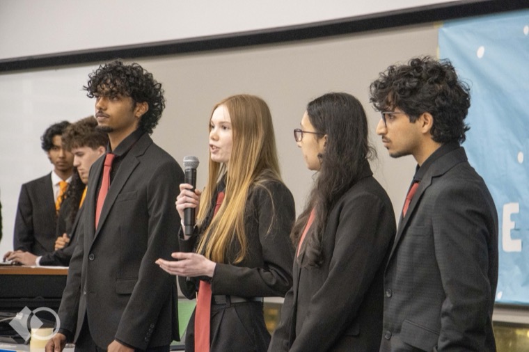 Madhav Suri presenting with his team at a competition, standing in a suit while a teammate speaks into a microphone
